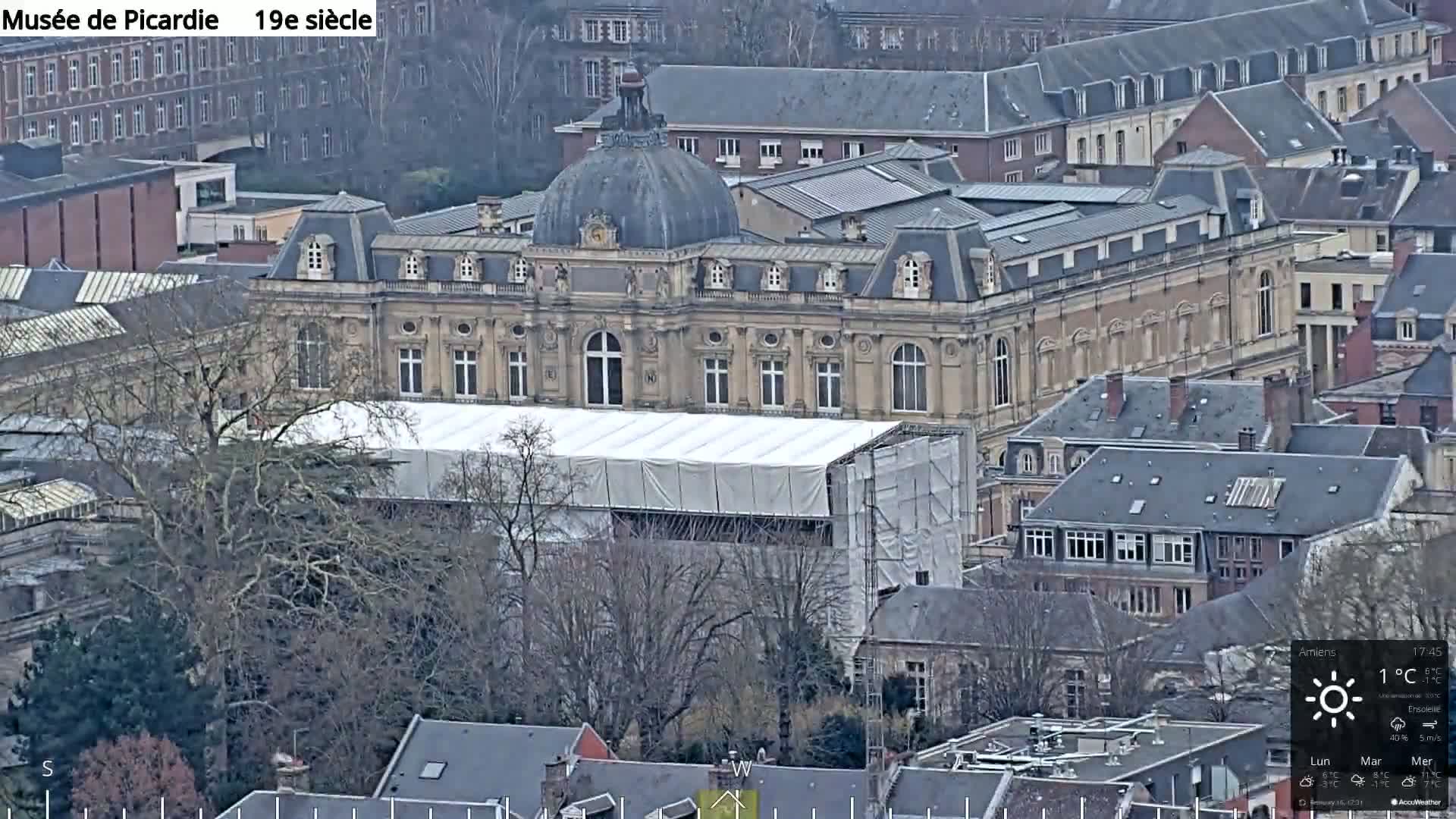 A large, tan building with a domed roof, partially covered by a white tarp and scaffolding, is seen from an elevated viewpoint on an overcast day.