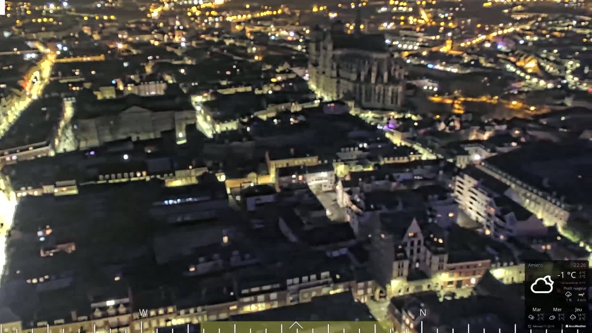 A nighttime aerial view of a city with many lights illuminating buildings and streets under partly cloudy skies.