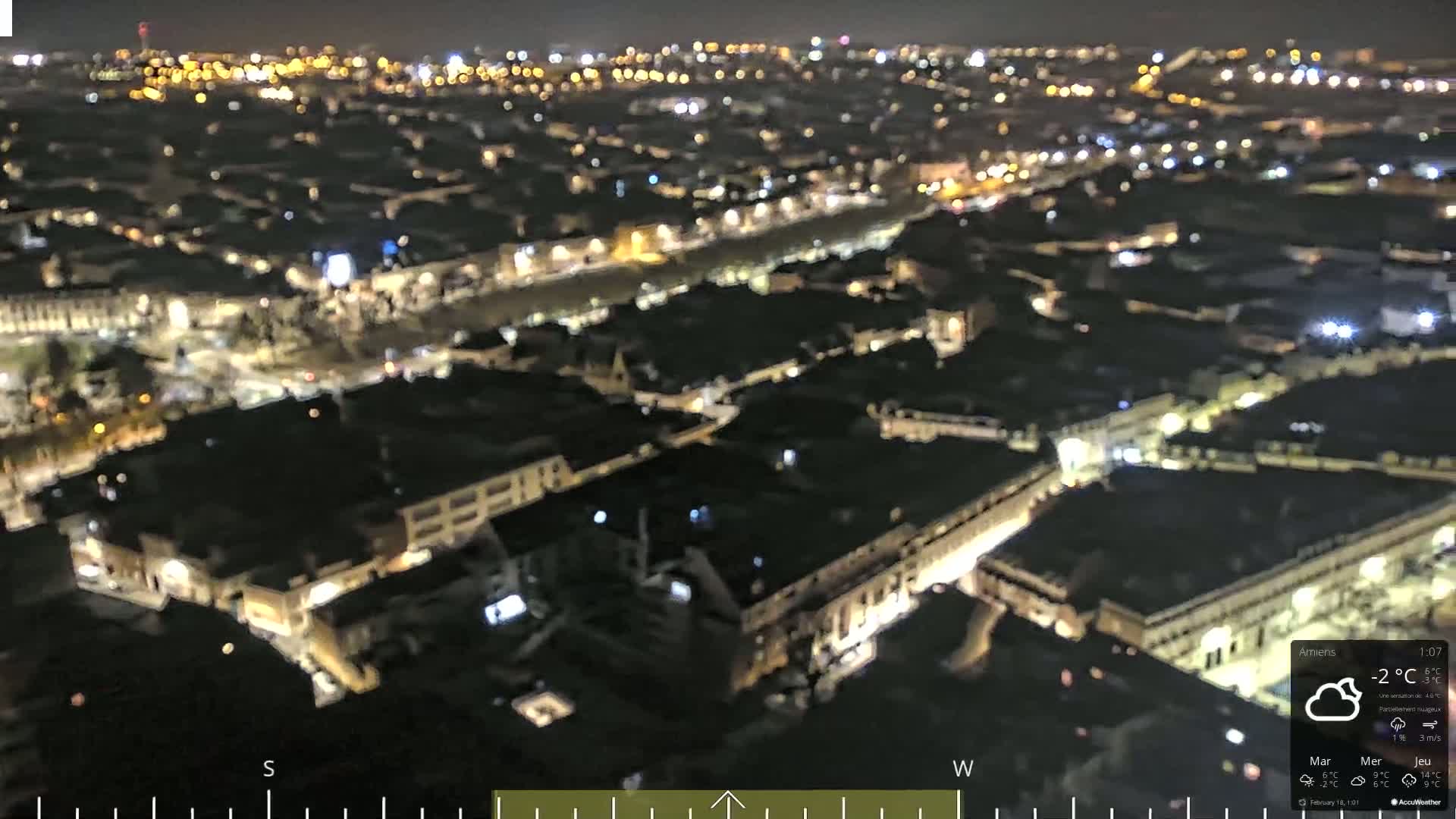A nighttime, high-angle, slightly blurry view of a city with many lights illuminating buildings and streets under partly cloudy skies.