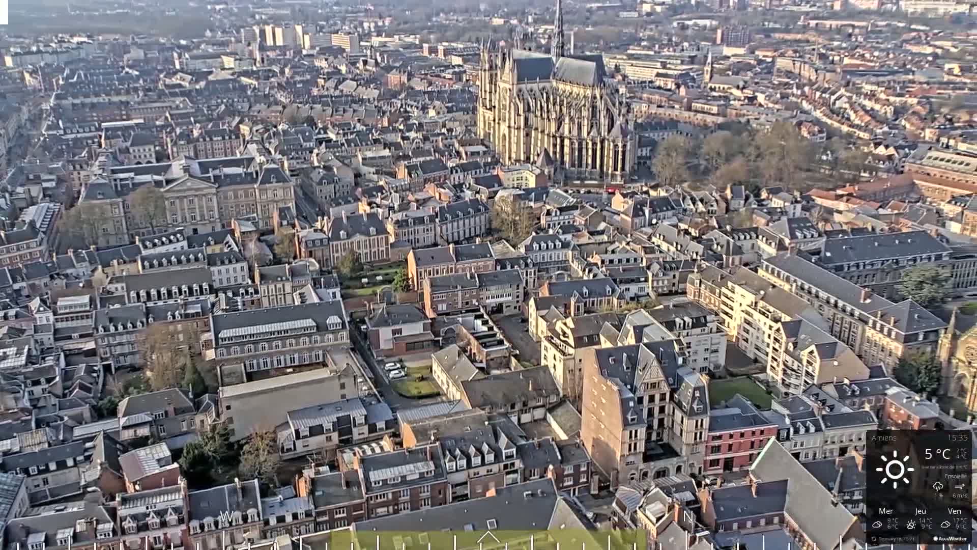 An aerial view of a densely populated city with a large cathedral in the center, under sunny skies.