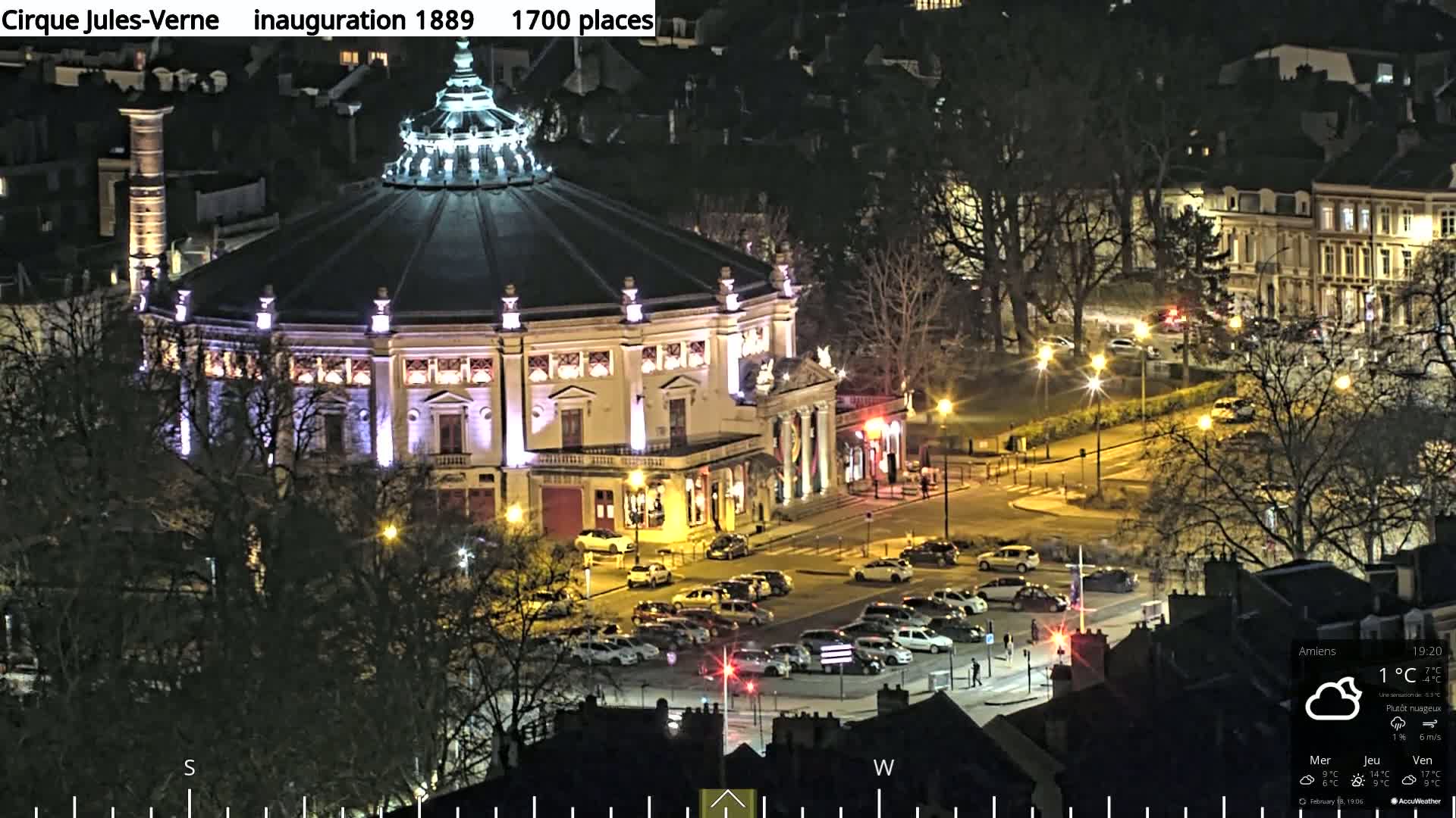 A large, round, illuminated building sits in a city square at night under partly cloudy skies.