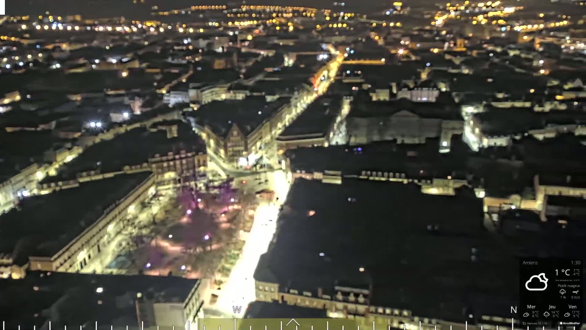 A nighttime aerial view of a city, illuminated by streetlights, under mostly cloudy skies.
