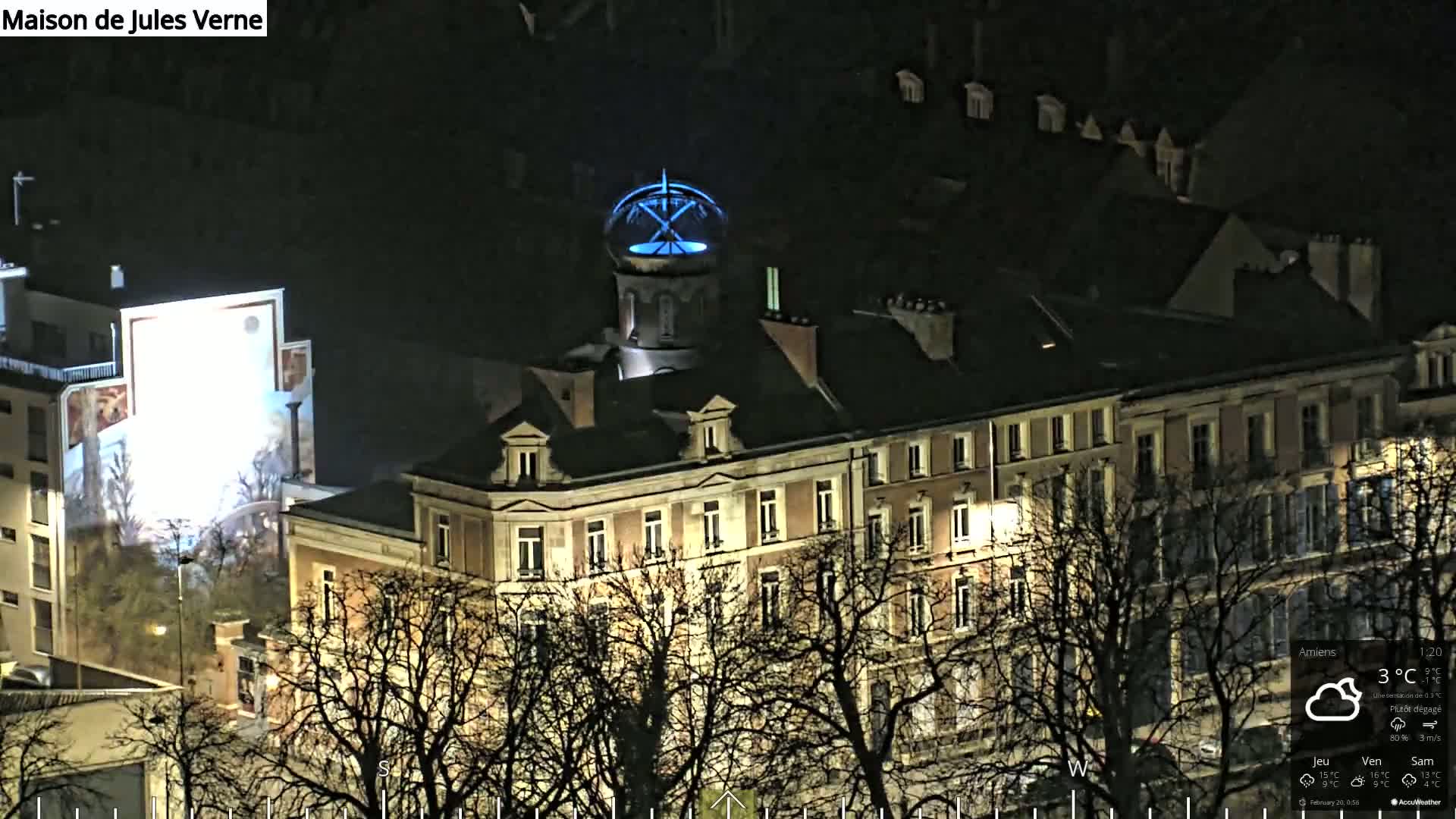 A nighttime view of a multi-story building with a cupola illuminated in blue, partially obscured by bare trees under a mostly clear sky.