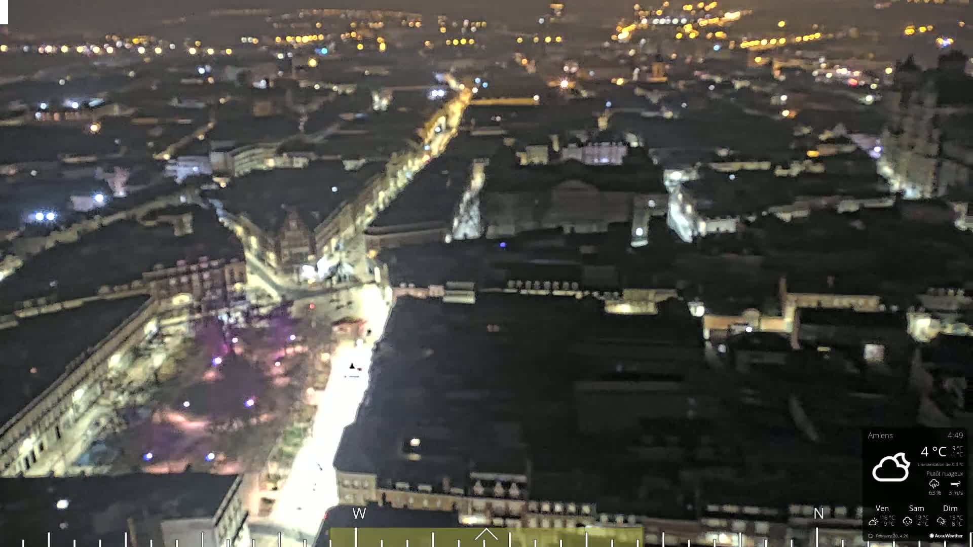 A nighttime aerial view of a city with a brightly lit main street, under partly cloudy skies.