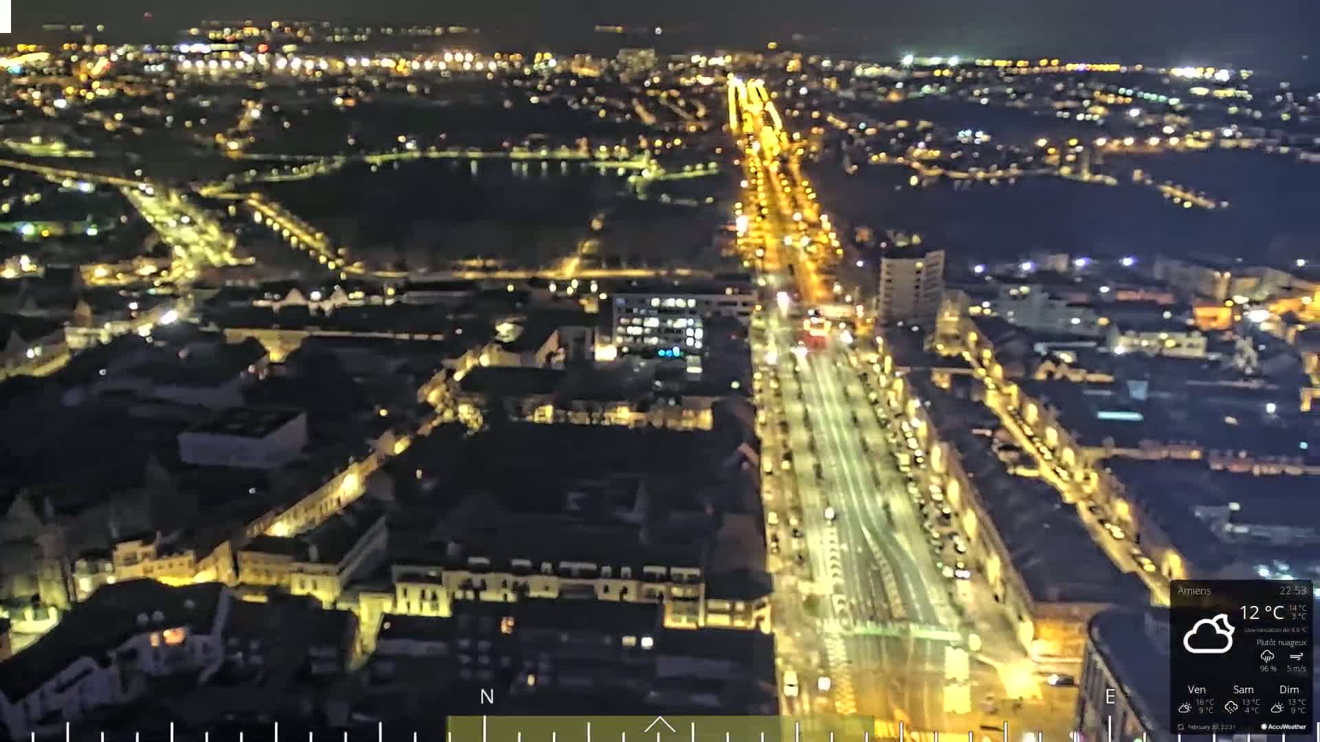 A nighttime aerial view of a city with brightly lit streets and buildings under partly cloudy skies.