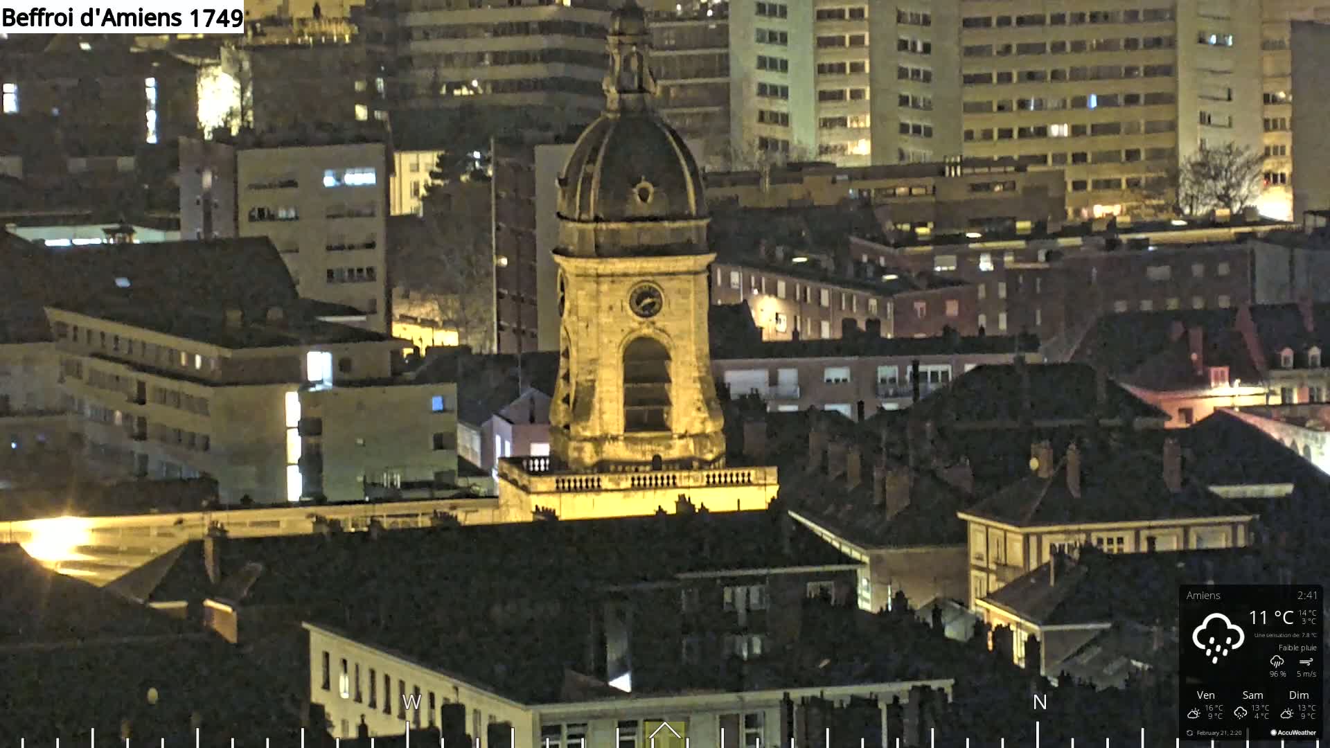A nighttime aerial view of a city, featuring a prominent clock tower, under a lightly rainy sky.