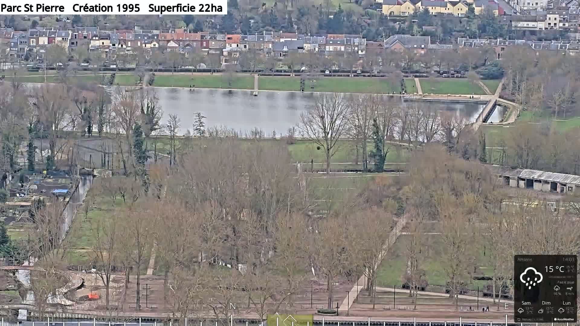 An aerial view shows a park with a large pond, bare winter trees, and pathways, under a slightly overcast sky with light rain.