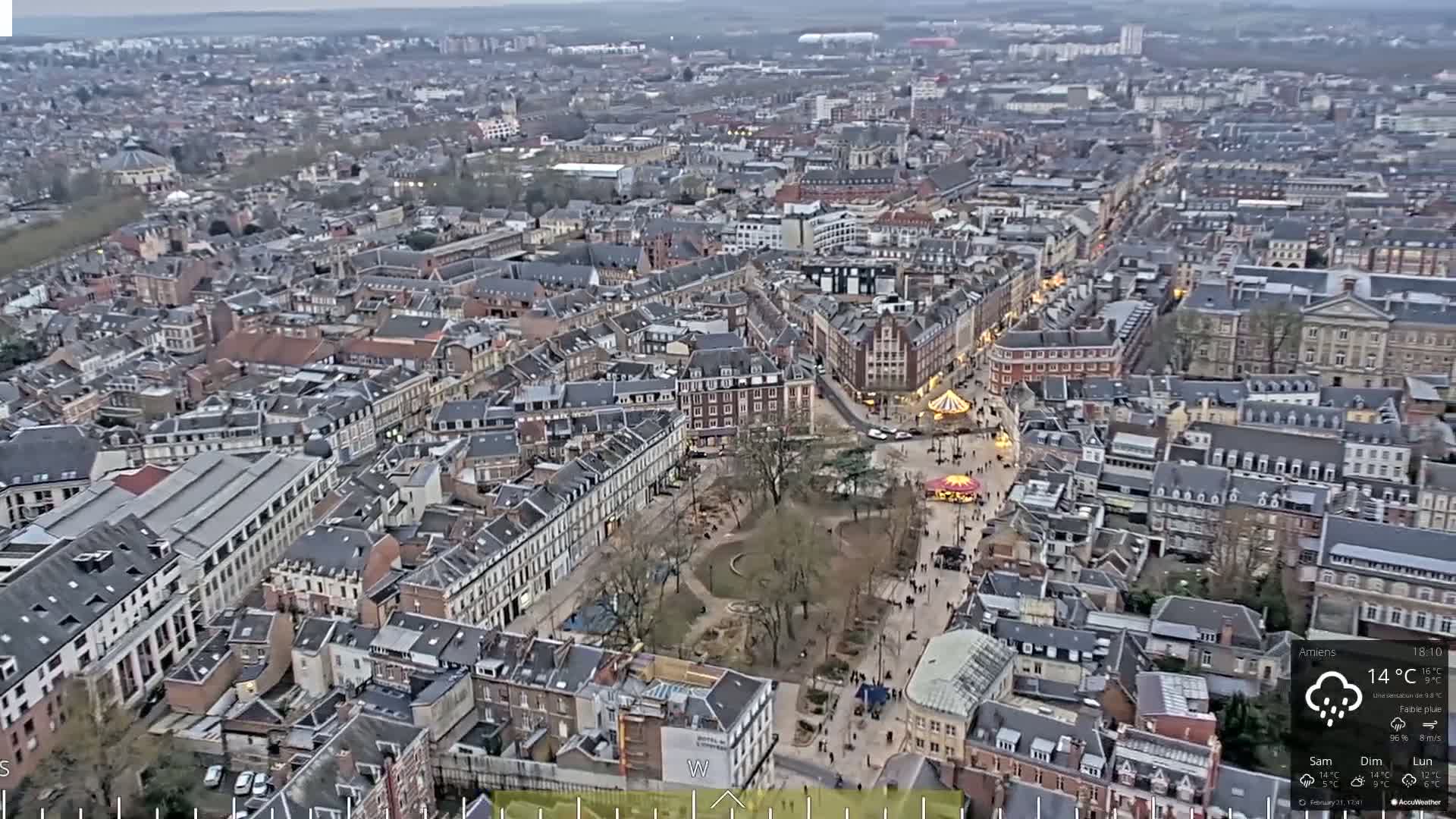 Here is a description of the image: An aerial view of a European city shows a large town square with a carousel and many pedestrians, surrounded by densely packed buildings under a slightly overcast sky with a light rain.