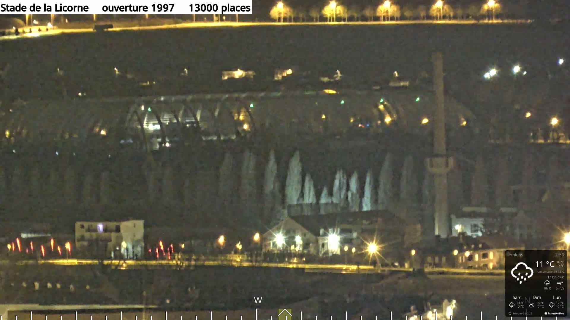 A nighttime view of a stadium with illuminated fountains and surrounding buildings under a slightly rainy sky.