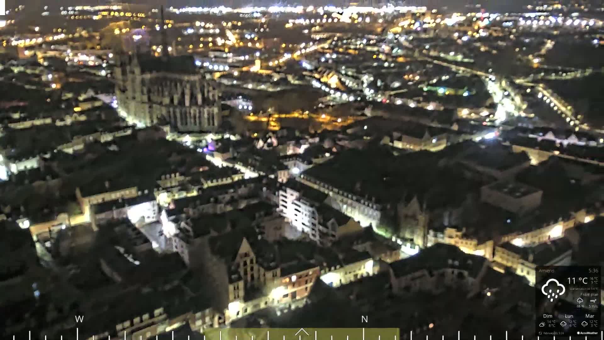 A nighttime aerial view of a European city with a large cathedral illuminated, under slightly cloudy conditions with light rain.