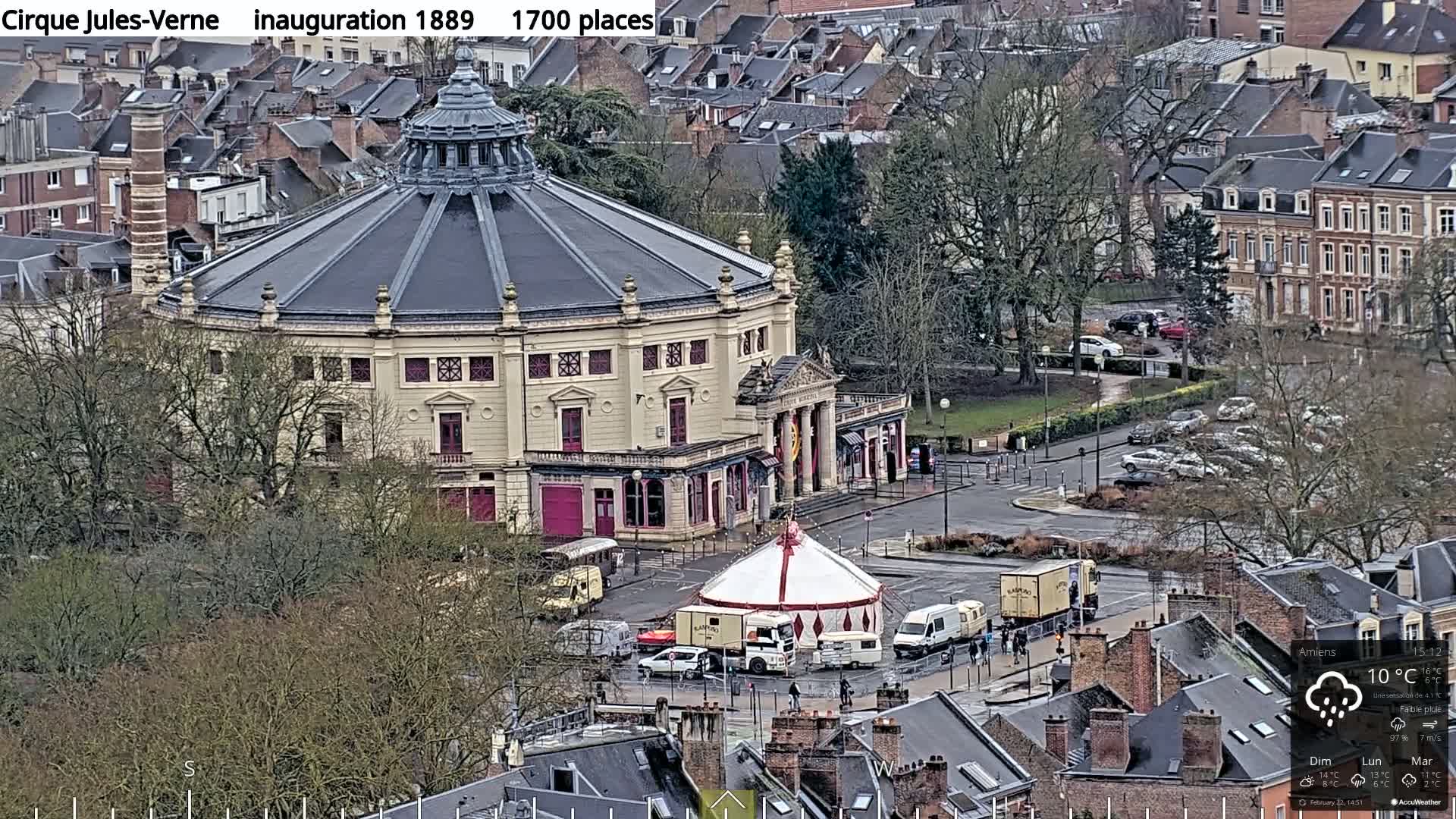 A large, round, light-beige building with a dark gray dome is surrounded by bare trees and parked cars in a city on a cloudy day with light rain.