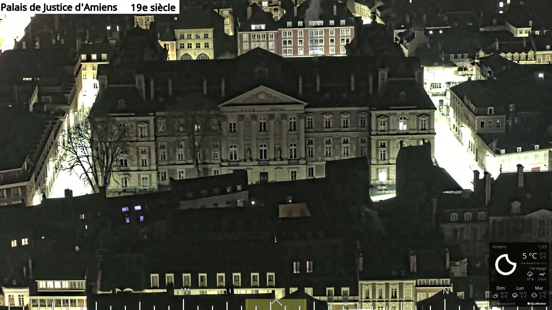 An aerial nighttime view of a city block with a large, brightly lit building in the center, surrounded by other dark buildings under a clear sky.