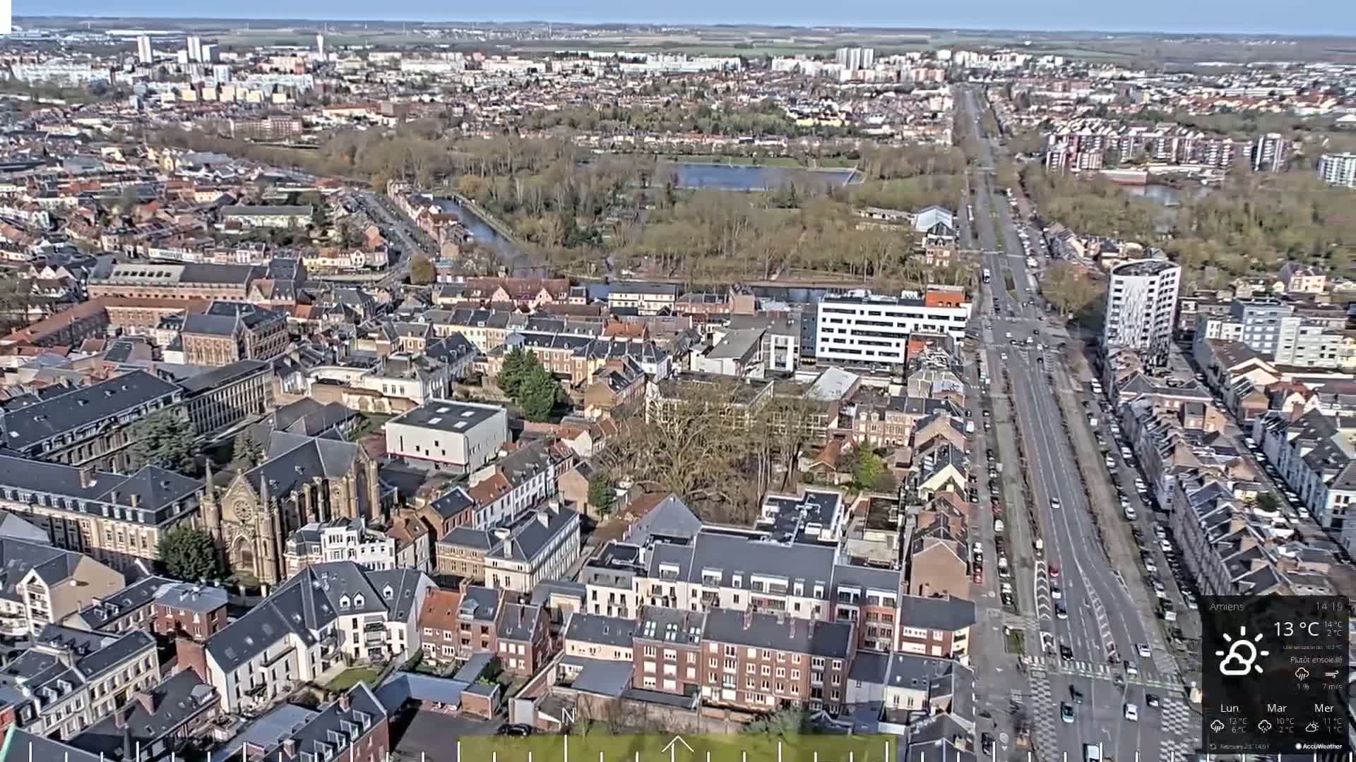 A high-angle, long shot of a city on a partly sunny day shows a network of streets and buildings, including a large church, a canal, and a wide road with parked cars.
