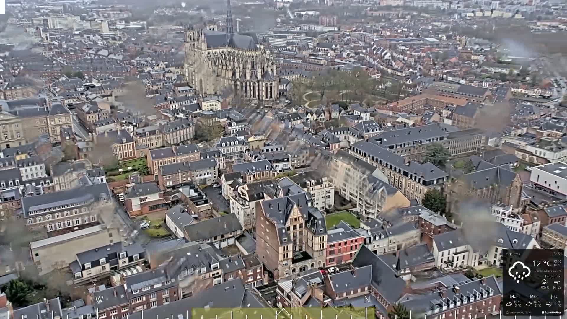 An aerial view of a European city on an overcast day with light rain shows a large cathedral and numerous densely packed buildings.