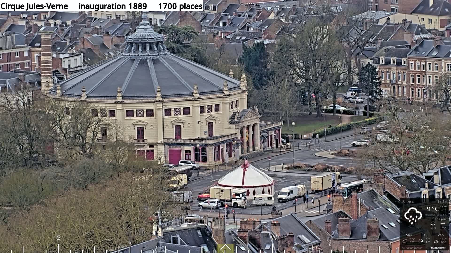 A large, round building with a dome-shaped roof sits in a city under a light rain, with a smaller, circus-style tent set up in front of it.
