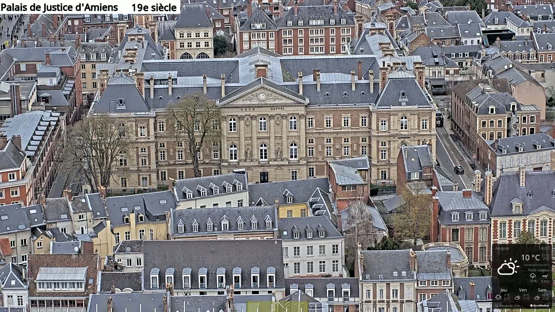 An aerial view shows a large, light-beige building with a dark gray roof surrounded by smaller, primarily gray-roofed buildings, under mostly cloudy skies.