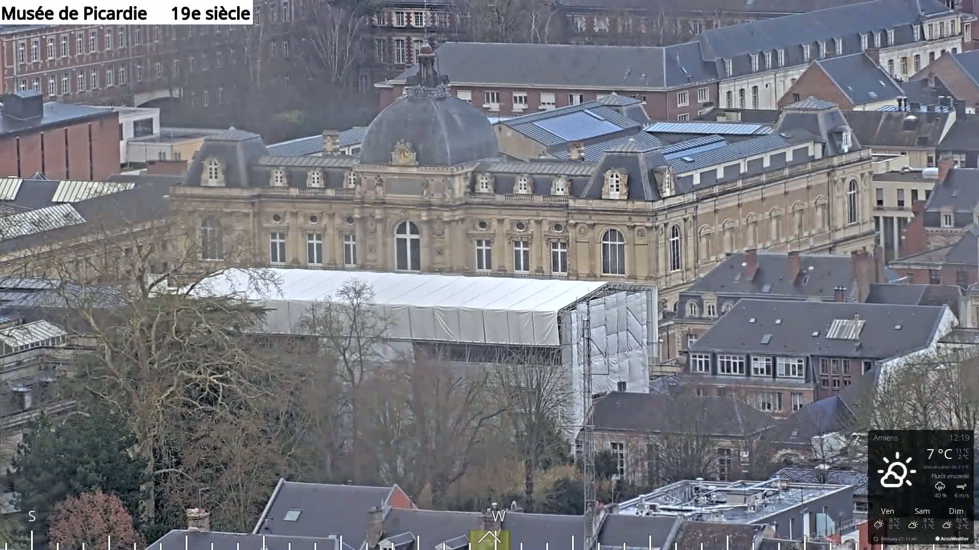 A large, light-brown building partially covered by a white tarp and scaffolding is surrounded by other buildings and bare trees under a mostly sunny sky.