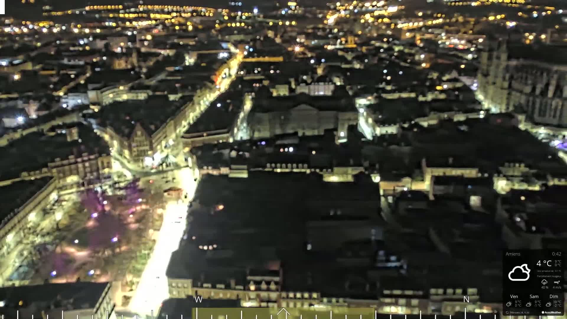 An aerial nighttime view of a city with dimly lit streets and buildings under cloudy skies.