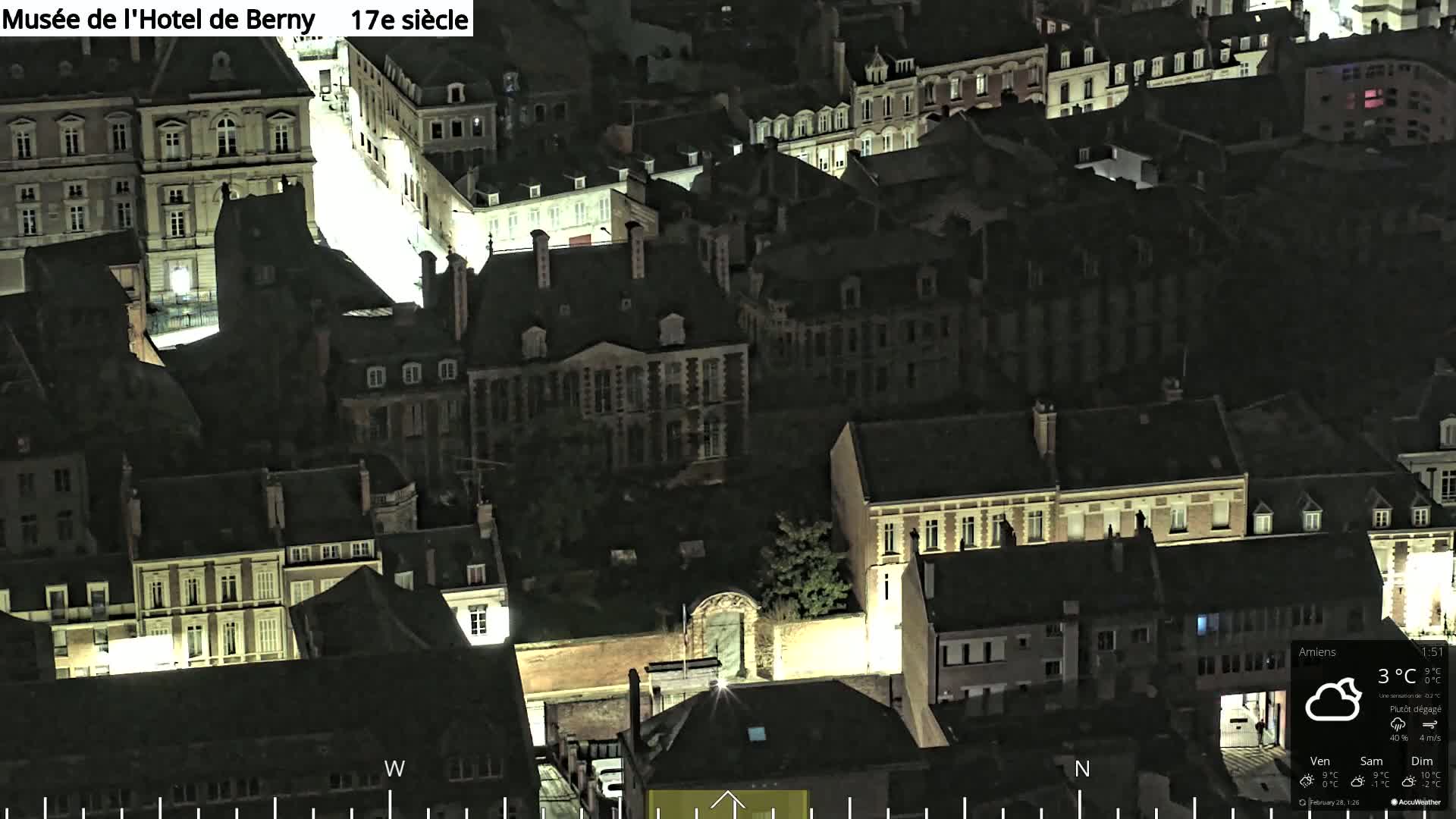 An aerial nighttime view of a city with numerous buildings illuminated, under partly clear skies.