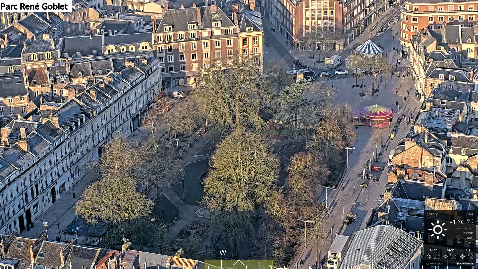An aerial view of a city square with a carousel, surrounded by buildings under a sunny sky.
