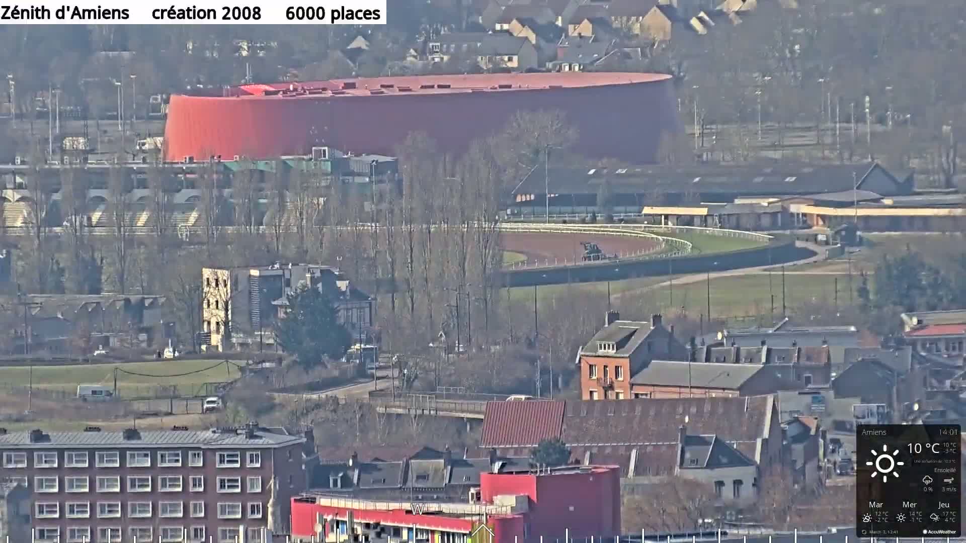A large red building sits beside a horse racing track in a town on a sunny day.
