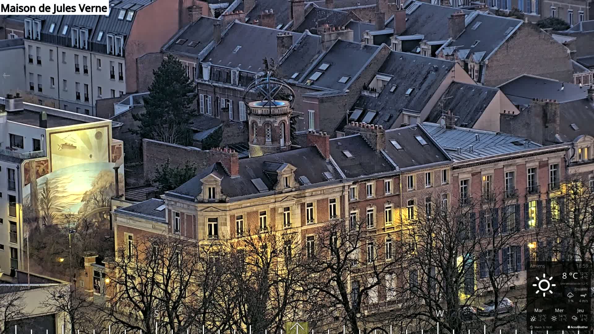 A nighttime aerial view of a city block with several buildings, including one with a mural and another with a distinctive tower, under a clear, sunny sky.