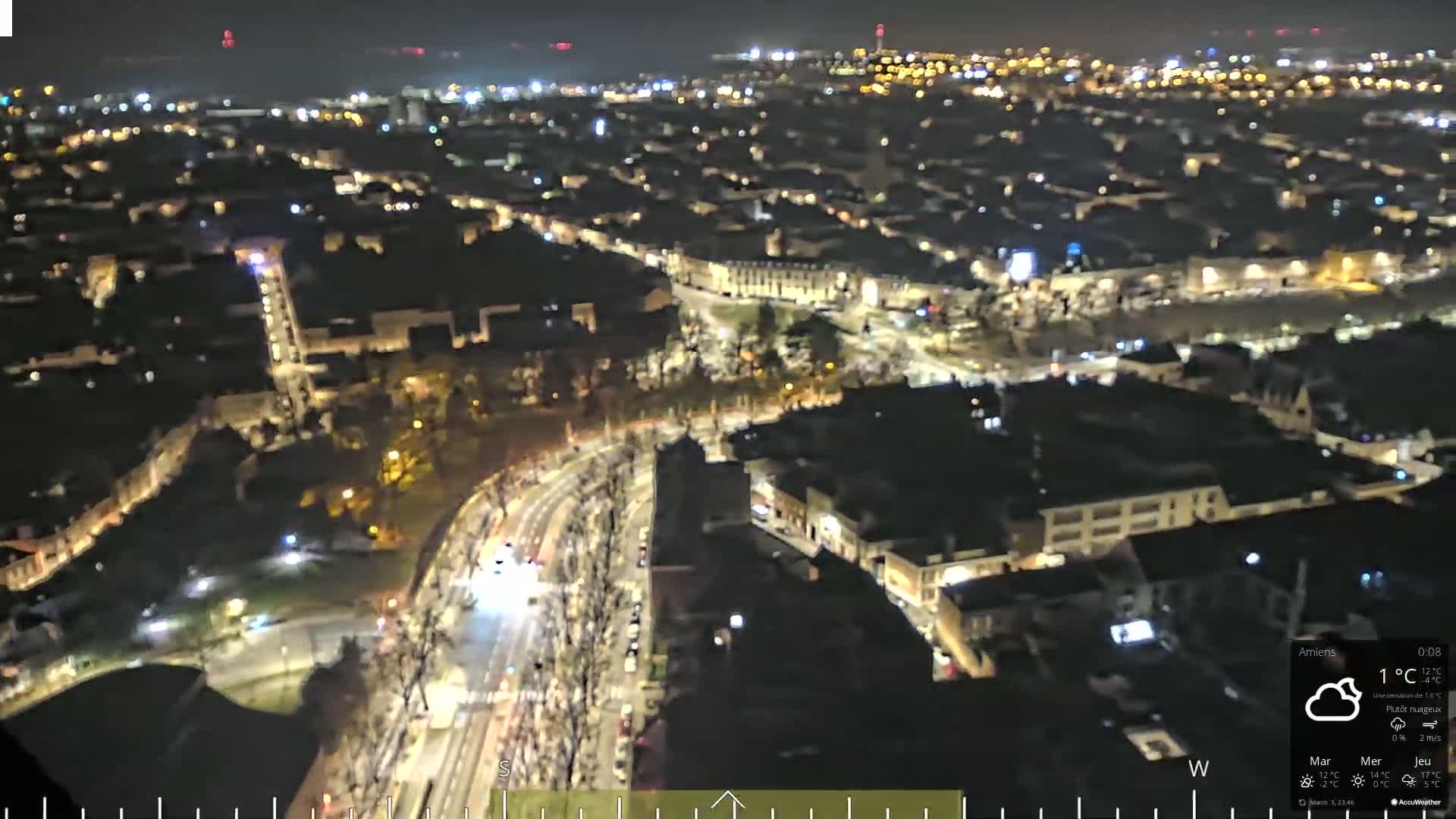 A nighttime aerial view of a city with illuminated buildings and streets, under partly cloudy skies.