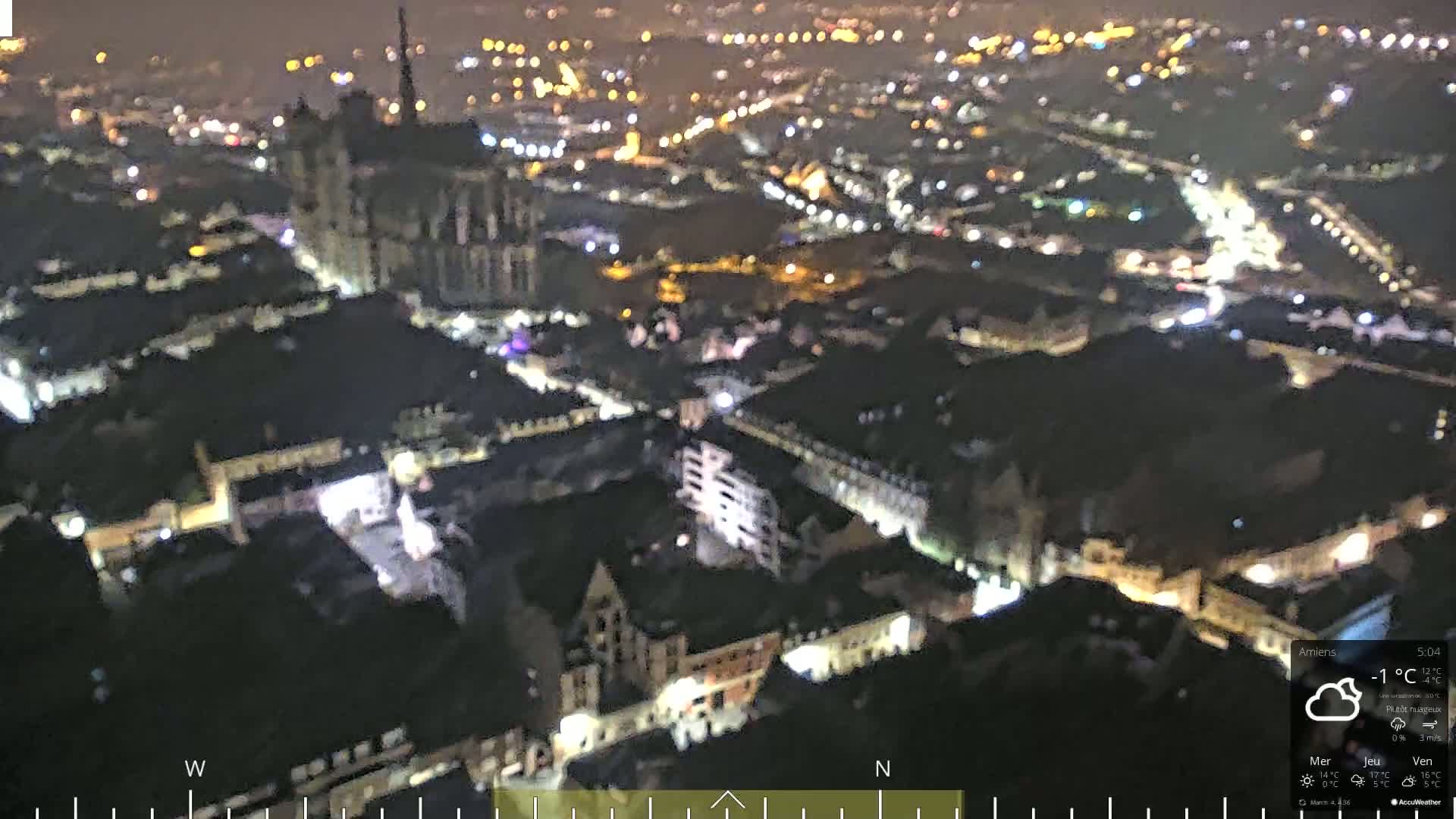 A nighttime aerial view of a city with a large cathedral, illuminated by streetlights under cloudy skies.