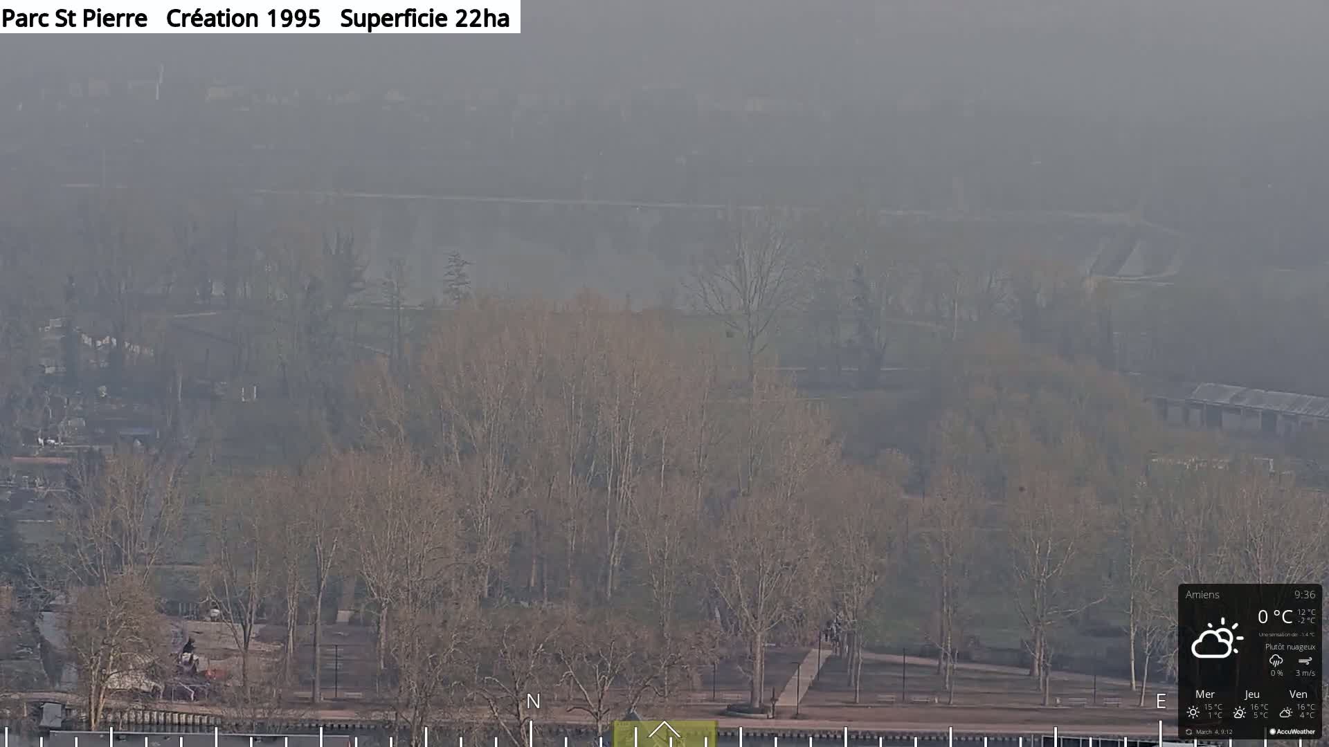 A hazy, mostly cloudy view of a park with bare trees and a body of water in the distance.
