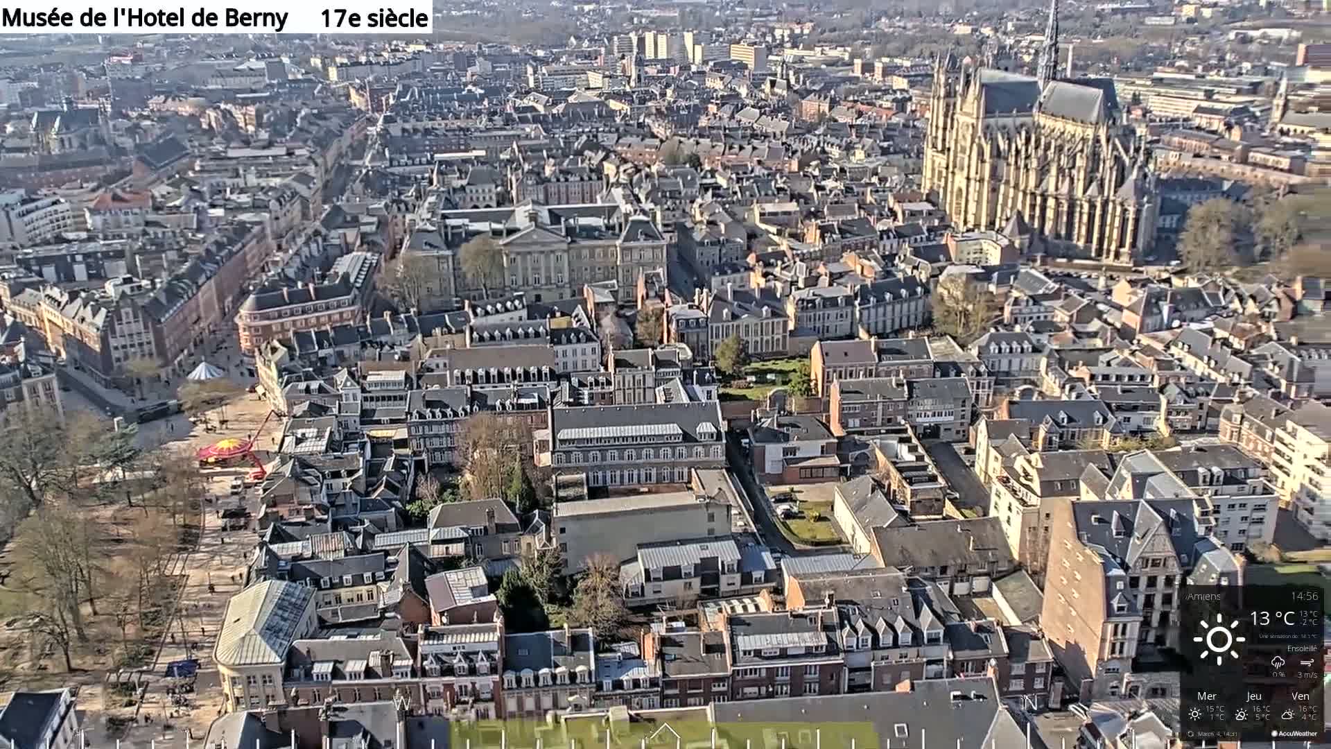 A high-angle, sunny view of a densely populated European city with a large cathedral, featuring a town square with a carousel and numerous buildings of varying heights and architectural styles.