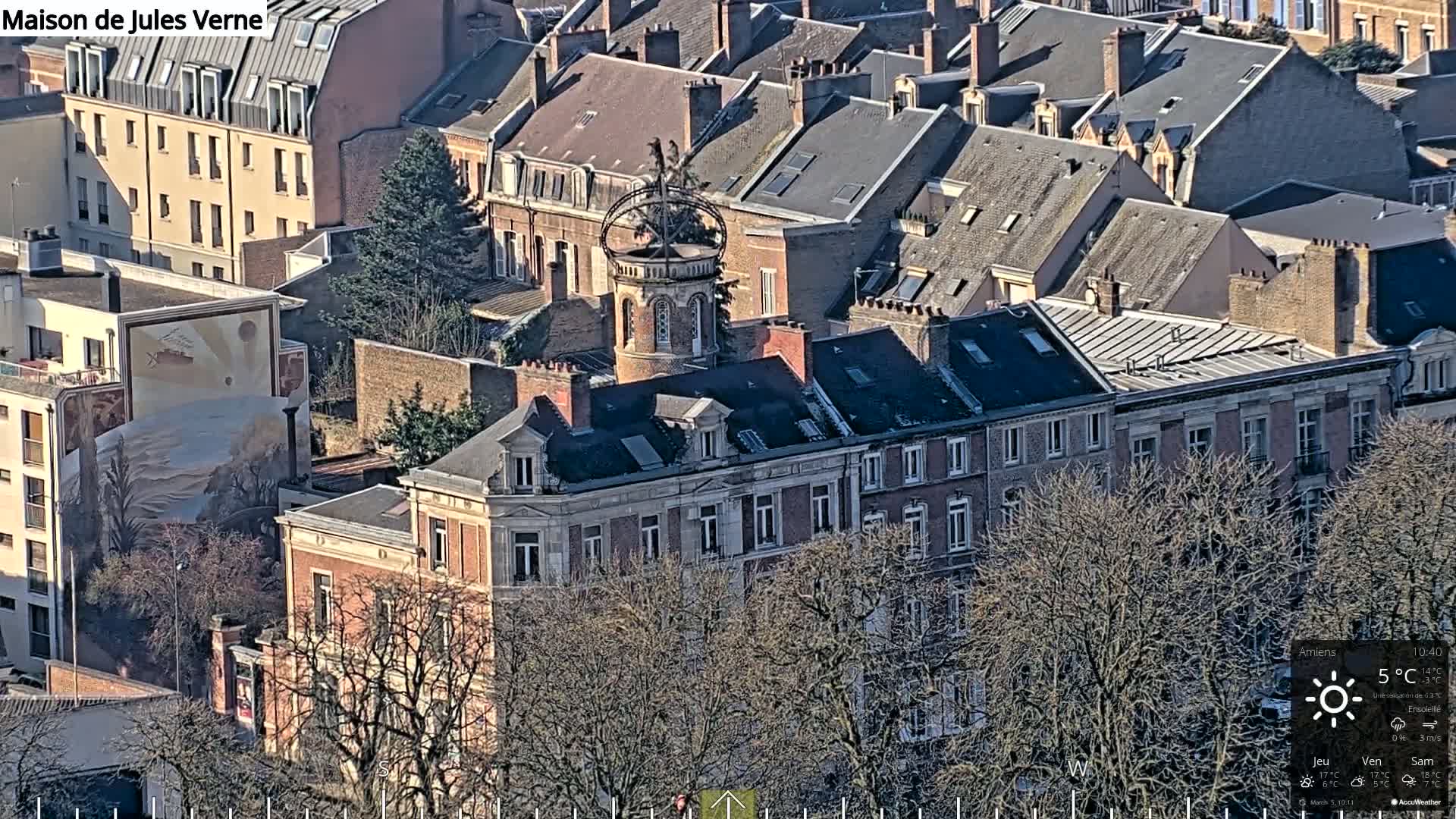 An aerial view shows a city block of closely spaced buildings with gray slate roofs, a central building featuring a tower with a decorative top, and leafless trees in a sunny, cold day.
