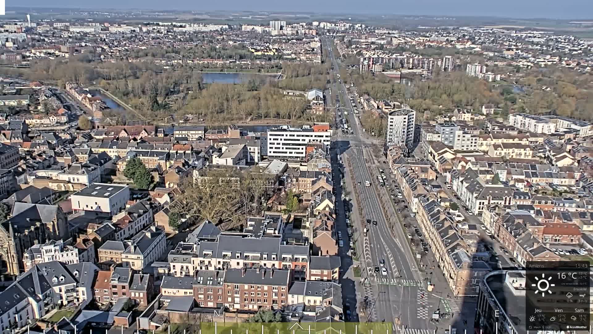 A high-angle, long shot of a city on a sunny day shows a main road cutting through residential areas, with a large park and body of water visible in the distance.