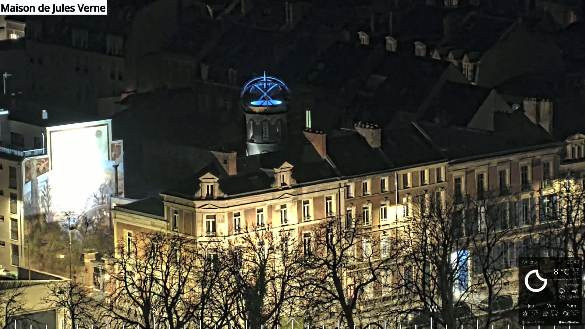 A large, multi-story building with illuminated windows is seen at night, partially obscured by bare trees under a clear sky.