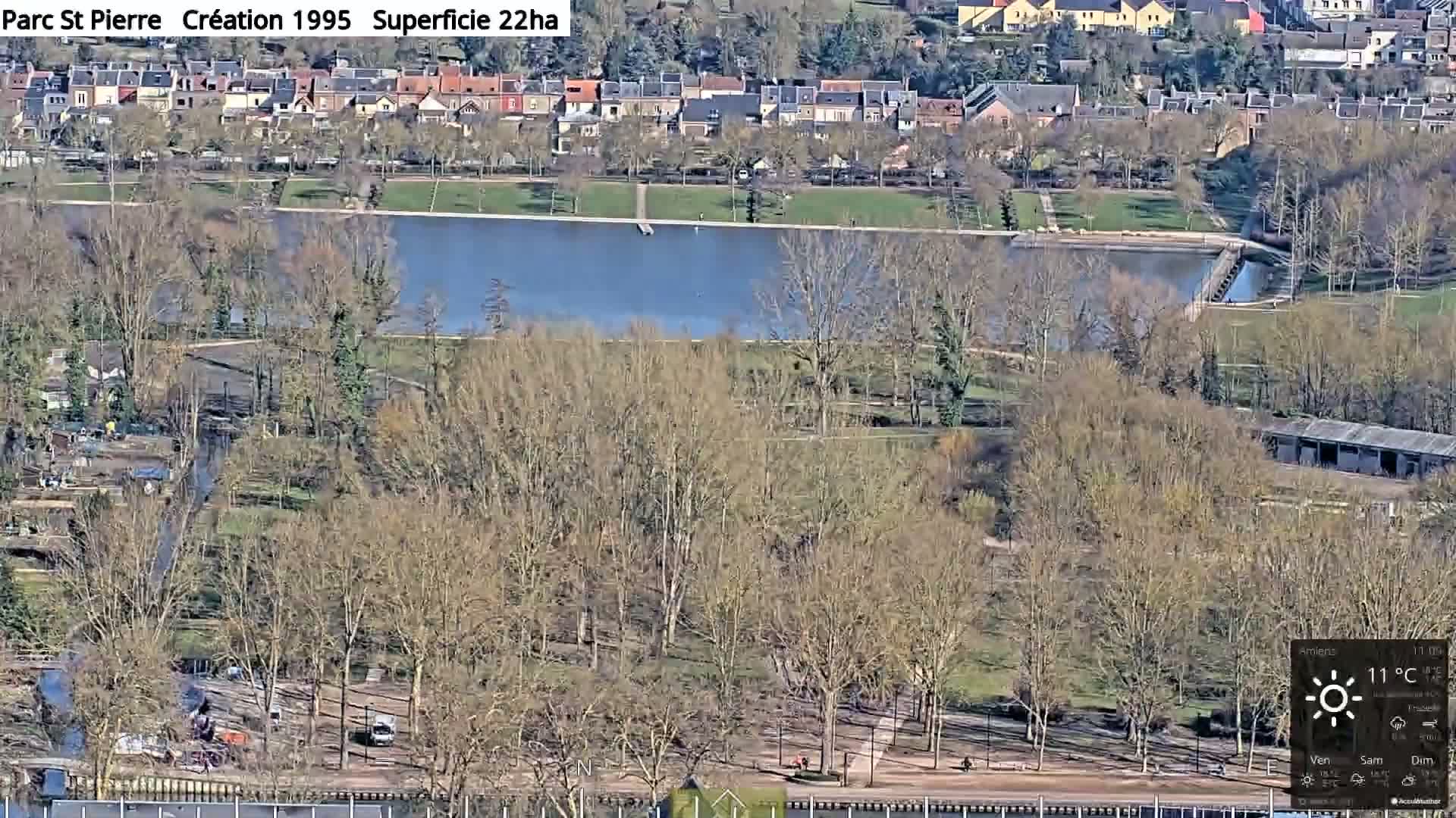 A high-angle view shows a park with a body of water, bare trees, and residential buildings in the background under a sunny sky.