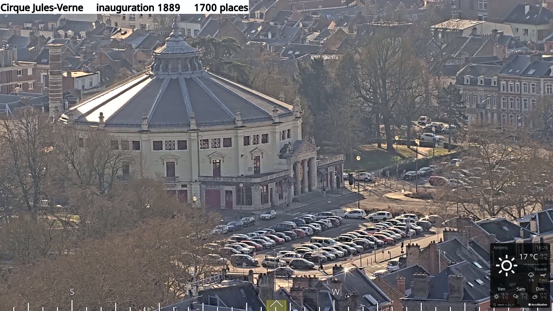 A large, round, white building with a dome-shaped roof sits in a city surrounded by bare trees and a parking lot full of cars under a sunny sky.