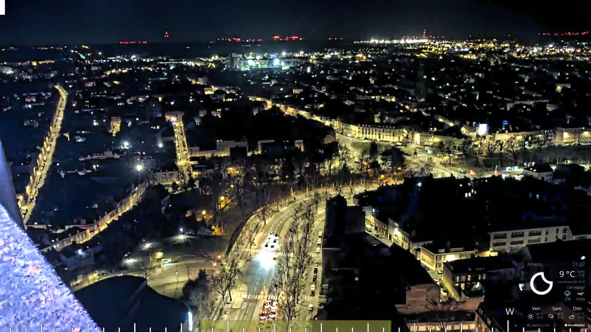 A nighttime aerial view of a city illuminated by streetlights, with a body of water visible in the distance under a clear sky.