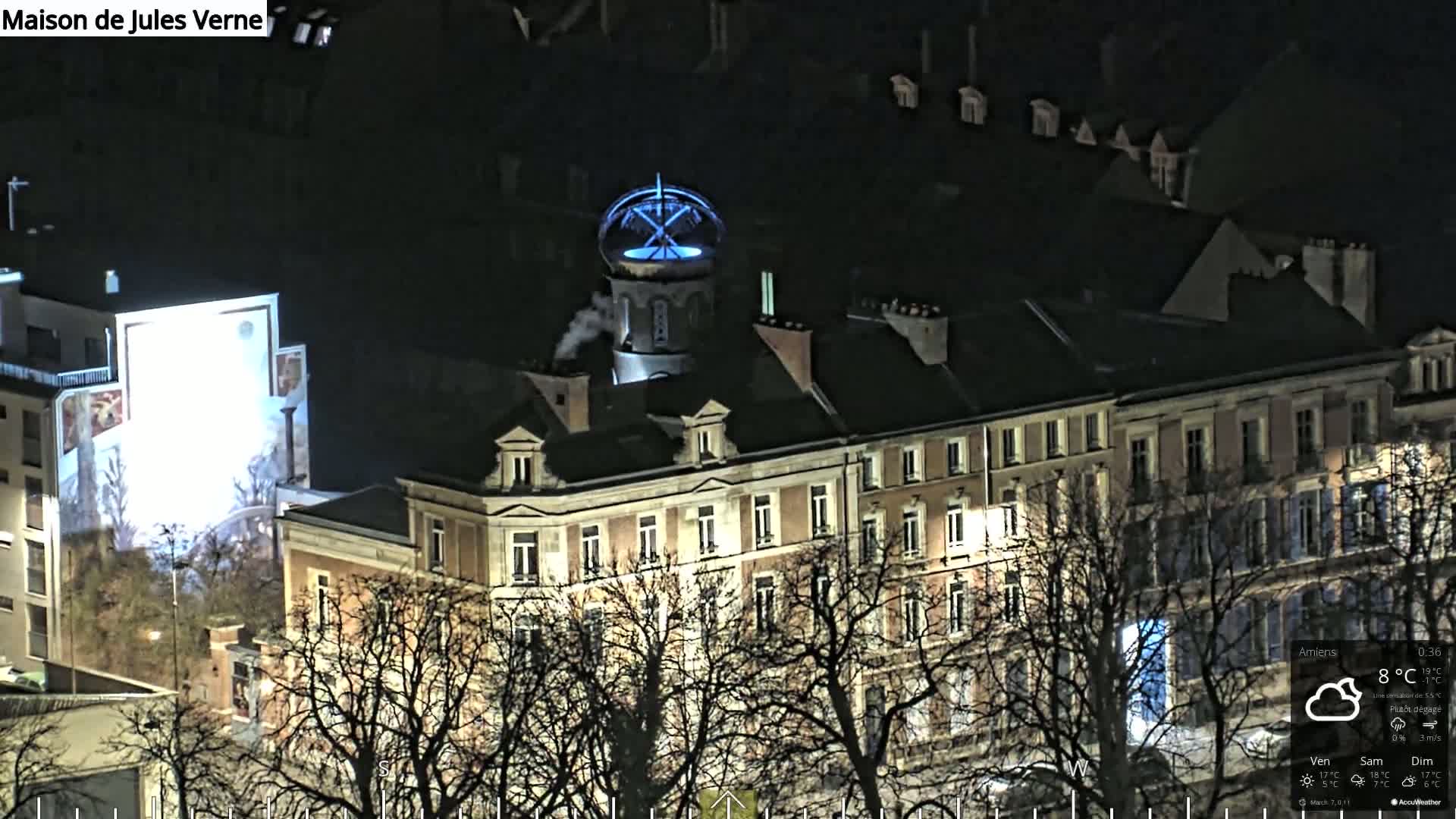 A large, multi-story building with a distinctive blue-lit cupola is illuminated at night, surrounded by bare trees under a mostly clear sky.