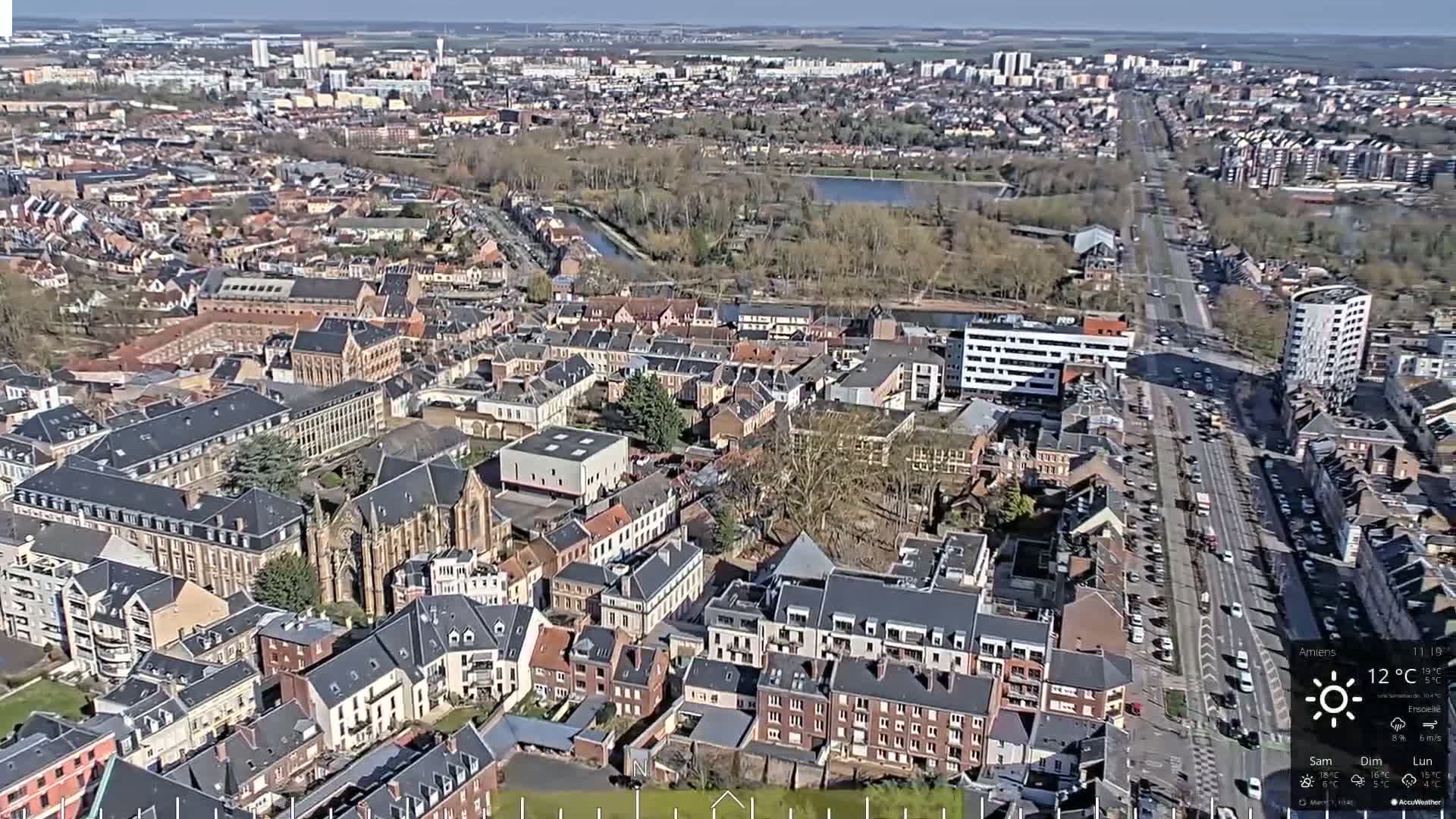 An aerial view of a city on a sunny day shows a dense collection of buildings surrounding a wide road, with a park and body of water visible in the distance.