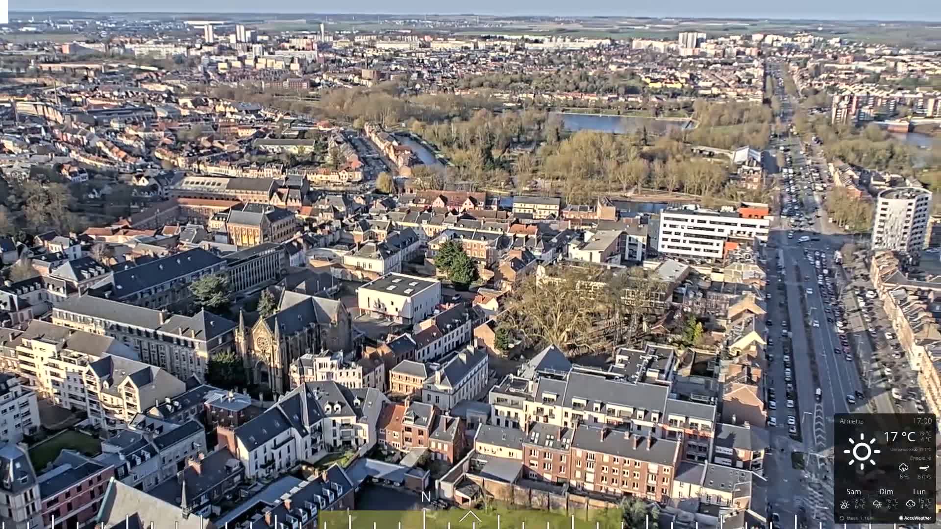 An aerial view of a European city on a sunny day shows a dense collection of buildings surrounding a long, straight road with moderate traffic.