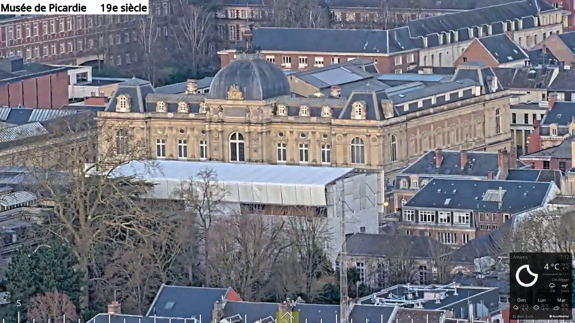 A large, light-brown building with a dome is partially covered by a white tarp, surrounded by other buildings on a cloudy day.