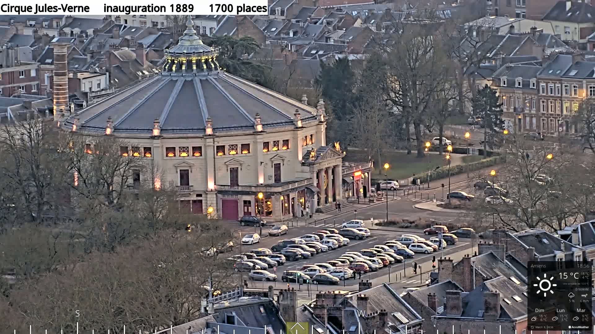 A large, round building with a dome-shaped roof and illuminated windows sits in a city at dusk, surrounded by parked cars and bare trees.
