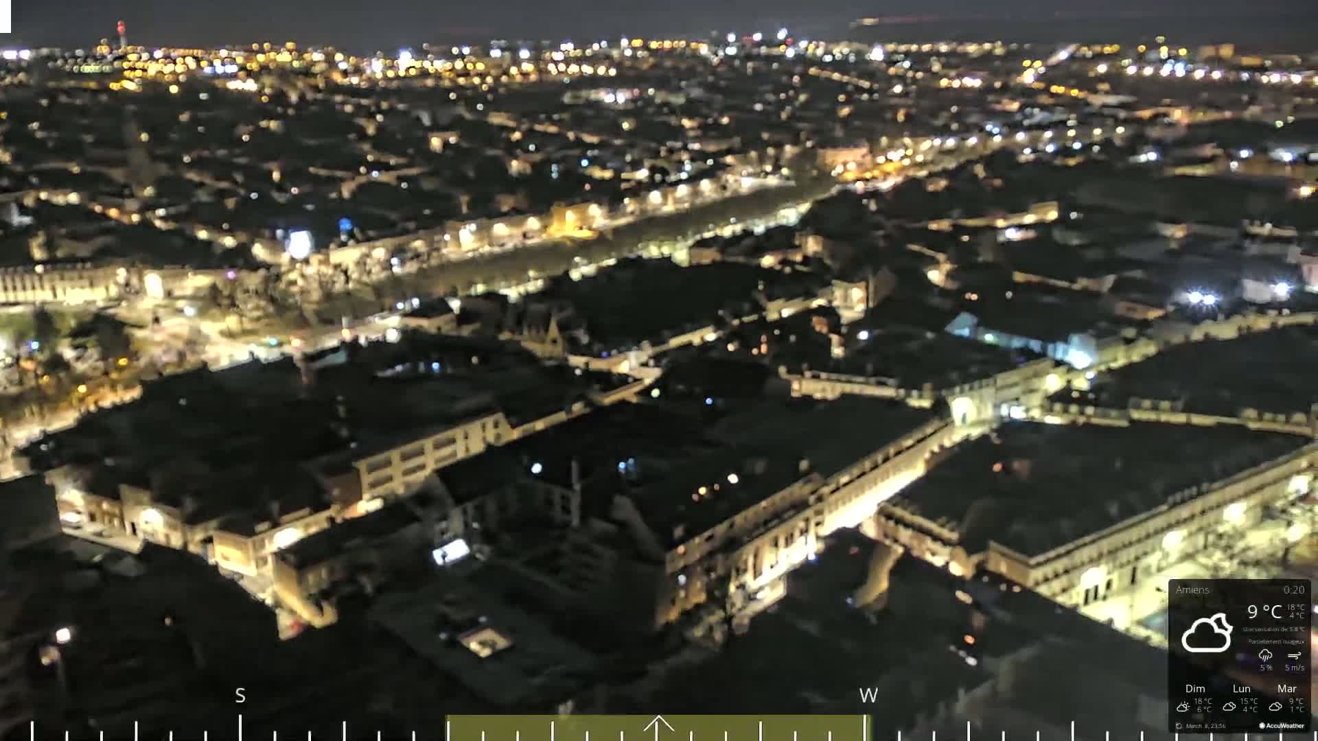 A nighttime aerial view of a city with numerous illuminated buildings and streets under a partly cloudy sky.