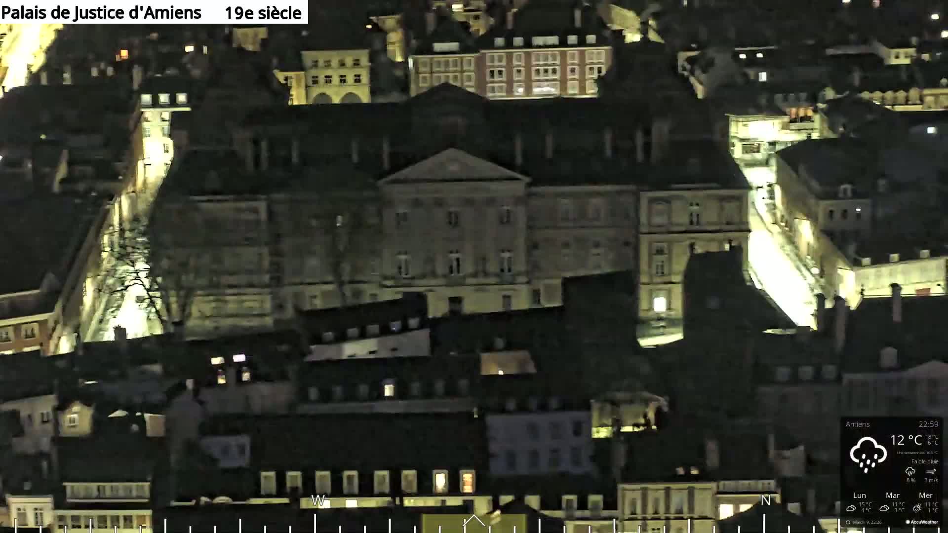 An aerial nighttime view of a city, showing a large building illuminated amidst a cluster of smaller, darker buildings under lightly raining conditions.