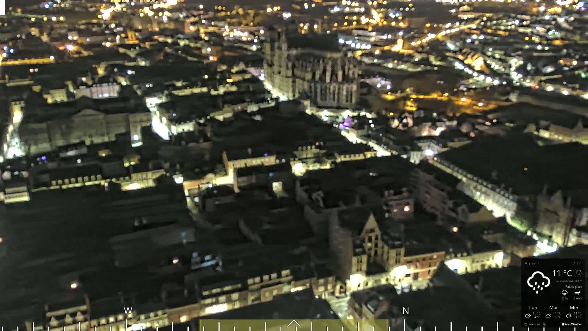 A nighttime aerial view of a city with a large cathedral at its center, under light rain.