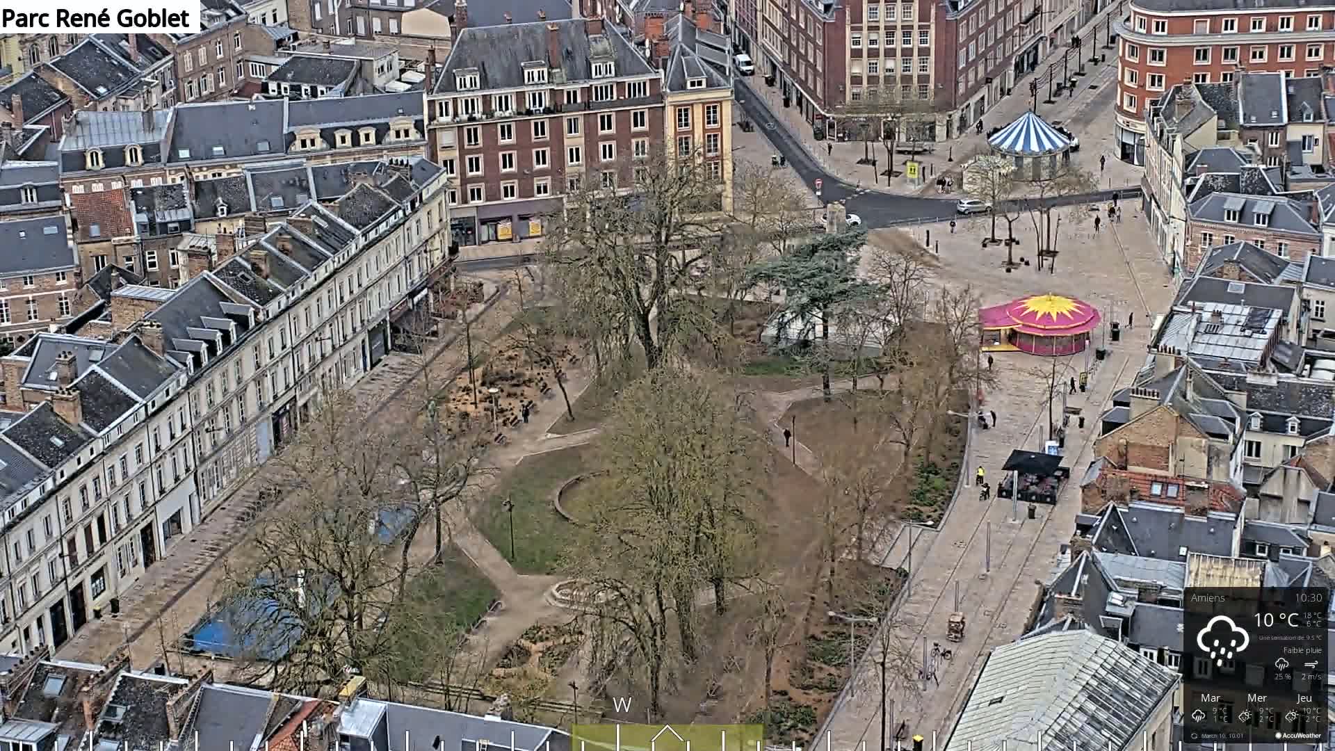 An aerial view of a city square with a park in the center, featuring a carousel and light rain.