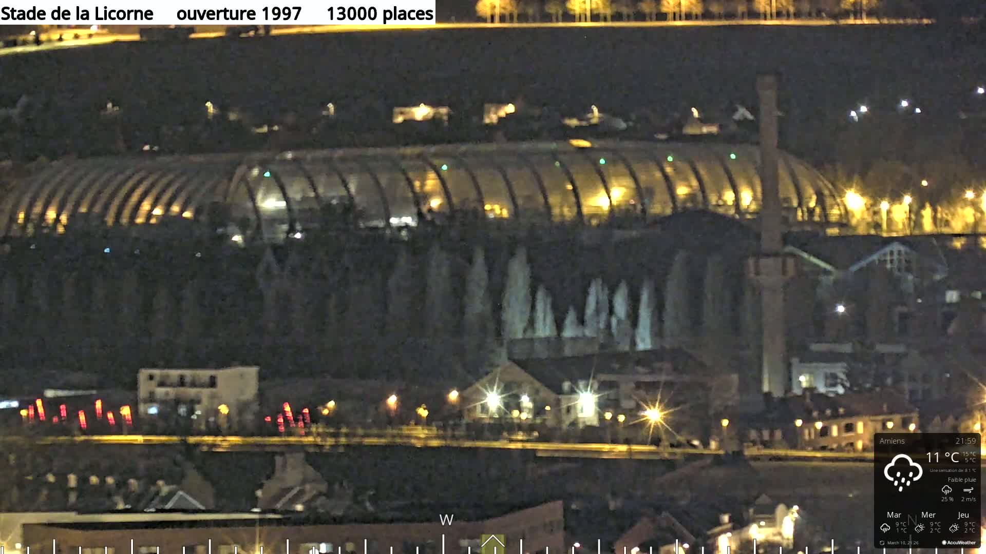 A nighttime view of a large, illuminated stadium with a curved roof, situated amongst smaller buildings and trees under light rain.