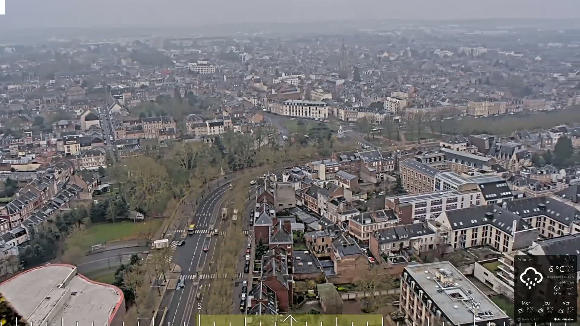 A high-angle, long shot of a city on an overcast day with light rain shows many residential buildings, roads, and some sparse trees.