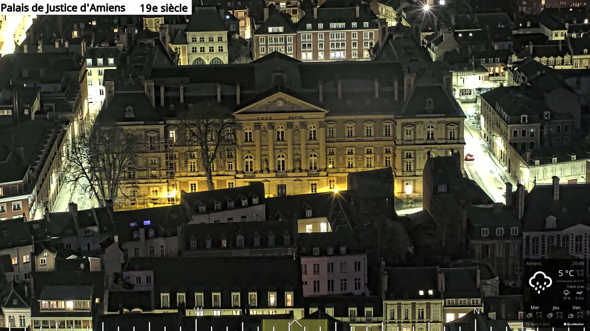 A large, illuminated building at night, surrounded by smaller, dark buildings under a light rain.