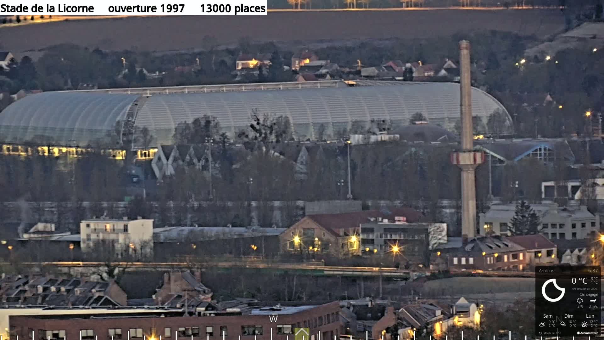 A large stadium with a curved, translucent roof is illuminated at night, nestled among residential buildings and a tall chimney, under a clear, dark sky.