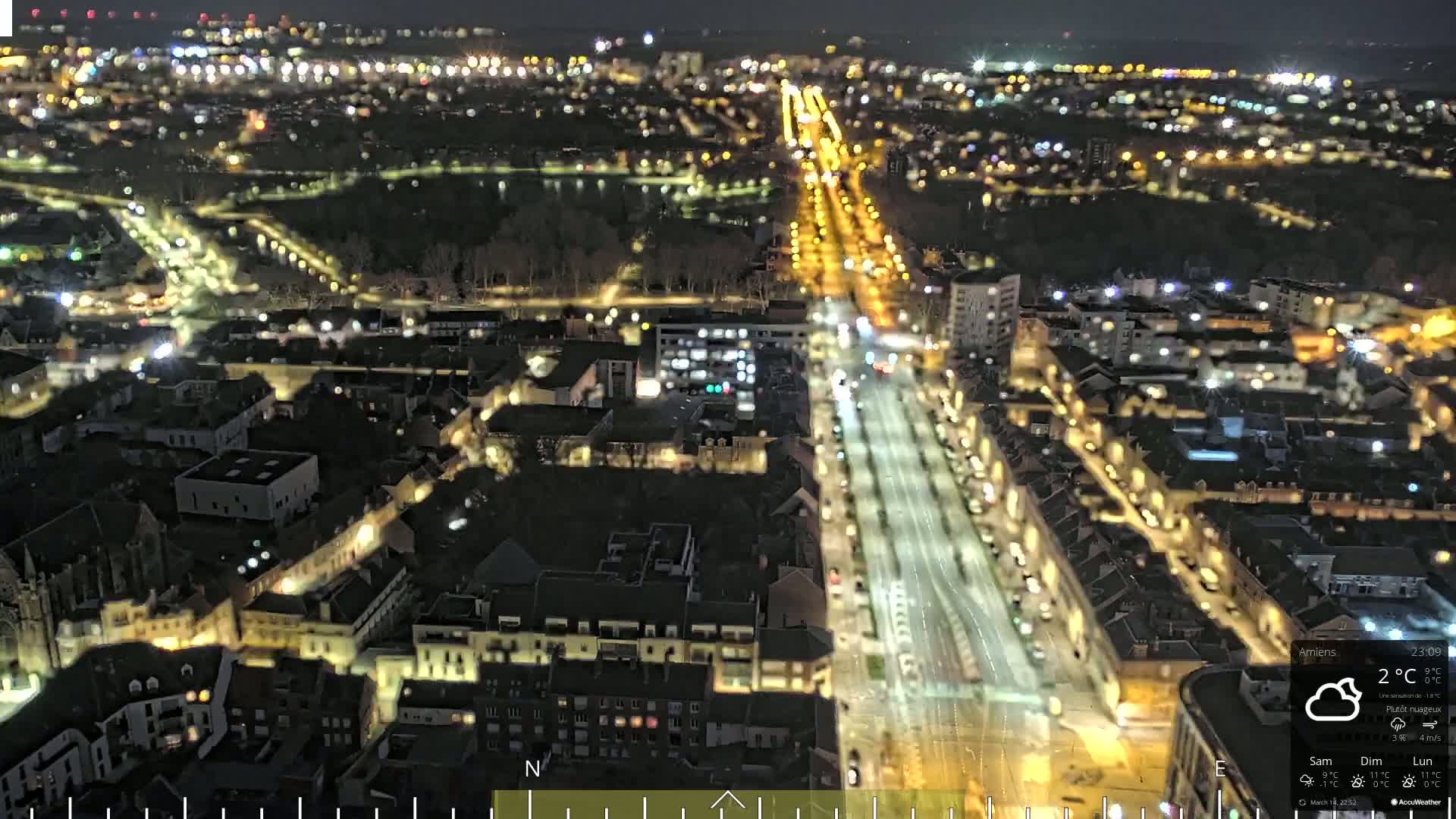 A nighttime aerial view of a city with brightly lit streets and buildings under partly cloudy skies.